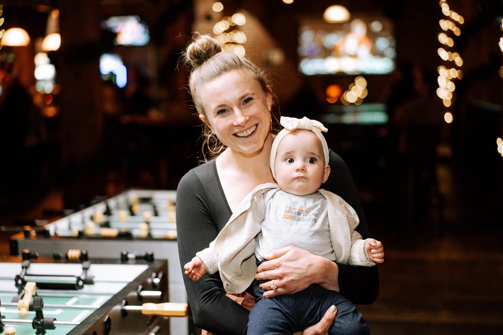 Woman smiles at the camera while holding her baby in front of foosball tables