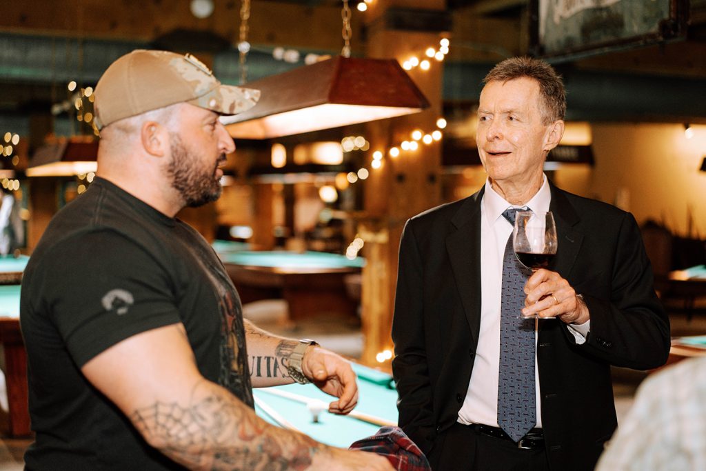 Man smiles and talks to another man while holding a wine glass