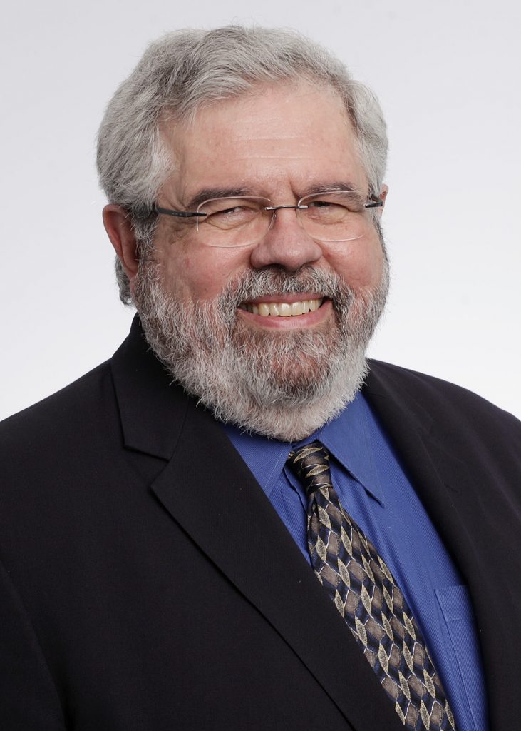 David Cay Johnston, a white man with short gray hair and gray beard, wearing a black suit jacket over a blue collared shirt, with a gray tie and thin, rectangular glasses, smiles.