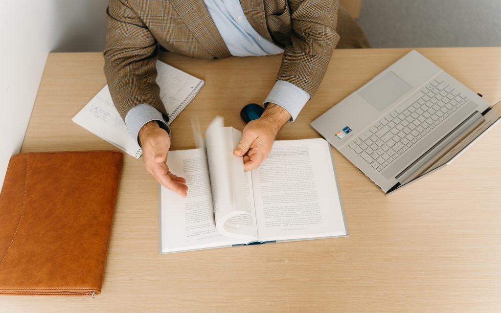 Abstract photo of man's hands leafing through a book and a laptop and leather briefcase on a desk in front of him.