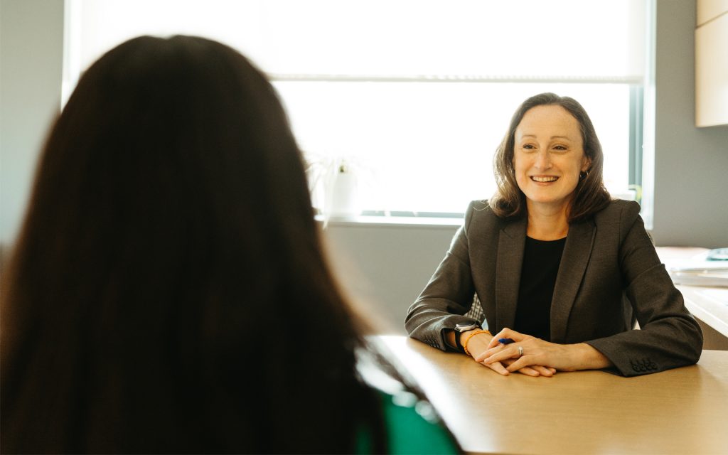 Professor Courtney Abbott Hill, in a gray suit, is focused as she writes on a document at her desk while a student in a green shirt watches attentively. Shelves filled with books and decor are visible in the background of the office.