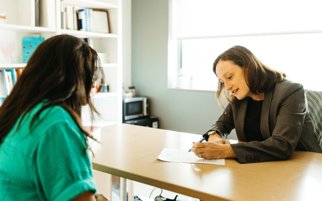 Professor Courtney Abbott Hill smiles warmly across a desk at a student during a one-on-one meeting. The student is seen from behind, wearing a green top, as they engage in conversation with Professor Abbott Hill in a brightly lit office.