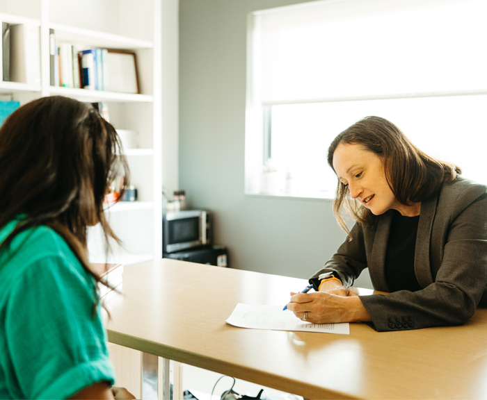 Professor Courtney Abbott Hill smiles warmly across a desk at a student during a one-on-one meeting. The student is seen from behind, wearing a green top, as they engage in conversation with Professor Abbott Hill in a brightly lit office.