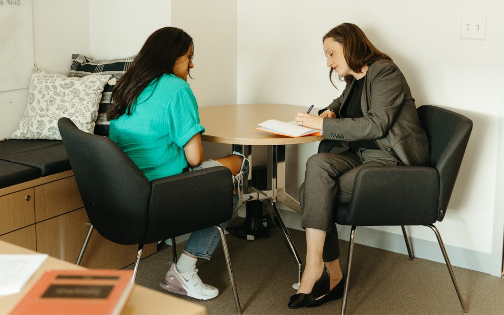 Professor Courtney Abbott Hill sits across from a student in a casual, supportive meeting setting at a round table. She is writing in a notebook while the student, dressed in a green top and jeans, listens intently.