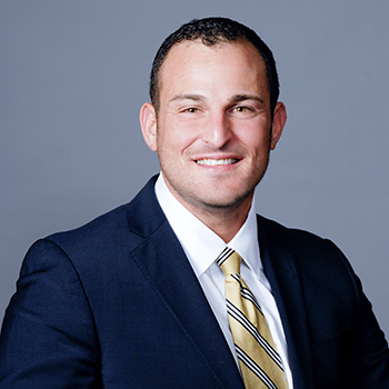 Adam Katz smiles at the camera for a headshot, wearing a blue suit in front of a plain blue background