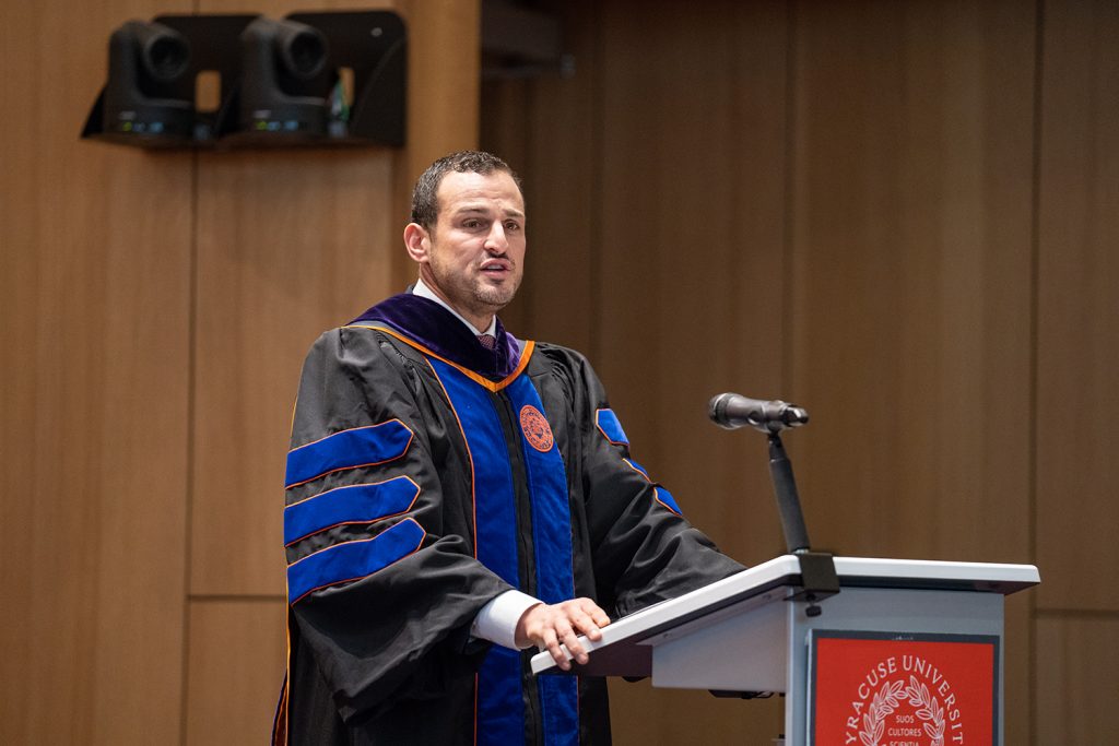 Adam Katz wears a robe as the keynote opening convocation speaker, speaking at a podium with a microphone