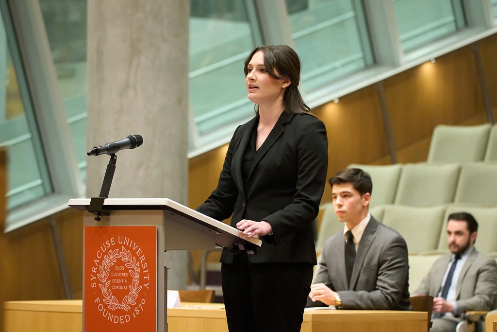 student stands at a podium speaking into a microphone in a courtroom with two students sitting behind her