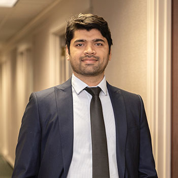 Ahmad smiles at the camera, standing in a hallway wearing a blue suit and a black tie