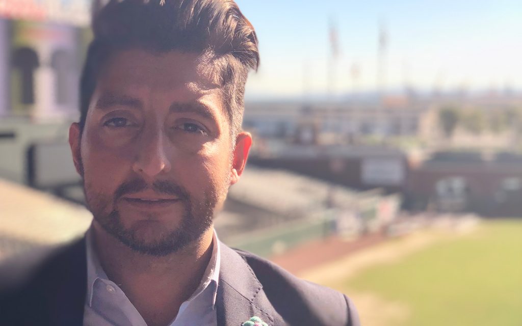 A man wearing a blazer standing at an outdoor location with blurred stadium seating in the background, looking at the camera.