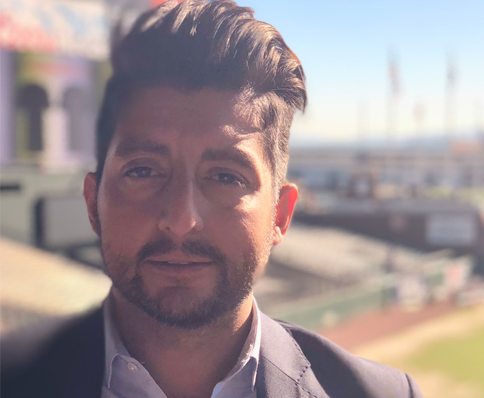 A man wearing a blazer standing at an outdoor location with blurred stadium seating in the background, looking at the camera.