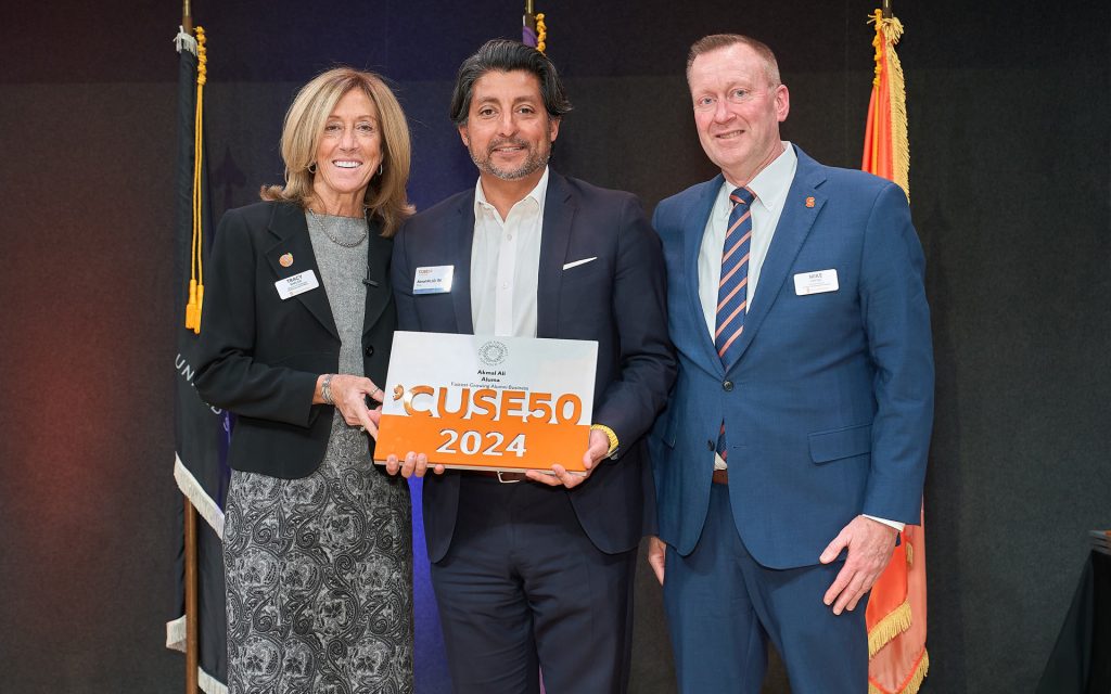 A man holding a 'CUSE50 2024 award plaque, standing between two other individuals in formal attire. They are smiling and posing in front of a dark background with flags.