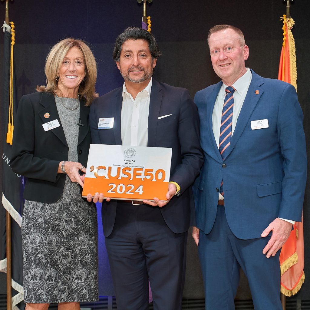 Three people smile holding a 'Cuse 50 sign up in front of a blue background and three flags.