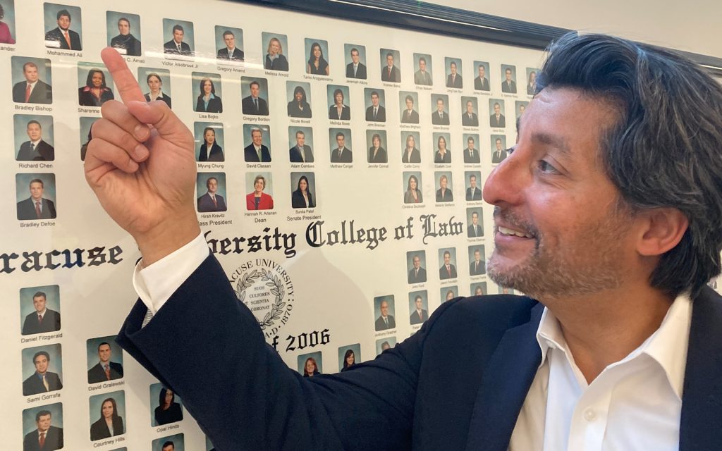  man smiling and pointing to his photo on a class composite portrait from Syracuse University College of Law, with a full view of his face and finger extended.