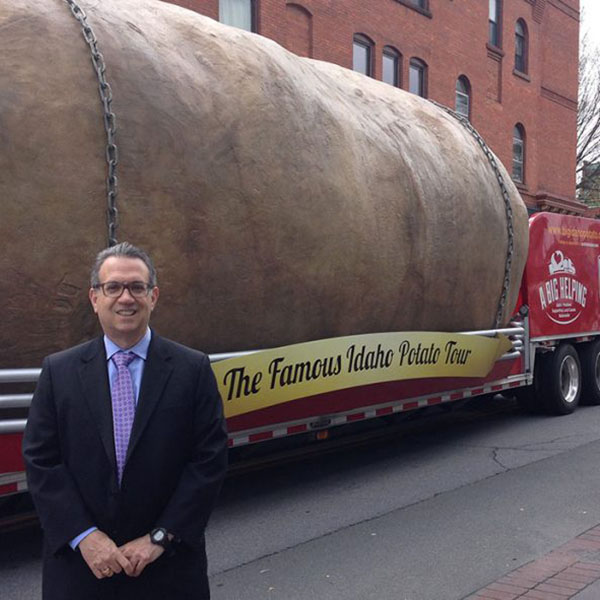 alan richter standing in a suit in front of a large potato on the back of an 18 wheeler truck