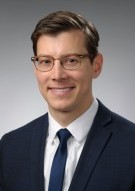 Andrew Horsfall, a white man with short brown hair, wearing a navy suit jacket over a white collared shirt with a navy tie, smiles in front of a white background.