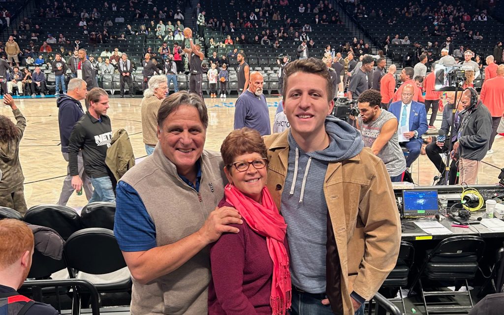 Zachary Mazuzan in a courtside photo at a Brooklyn Nets game smiling with his parents.