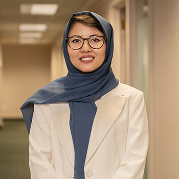 Beheshta smiles at the camera standing in a hallway, wearing a blue hijab and a white suit jacket