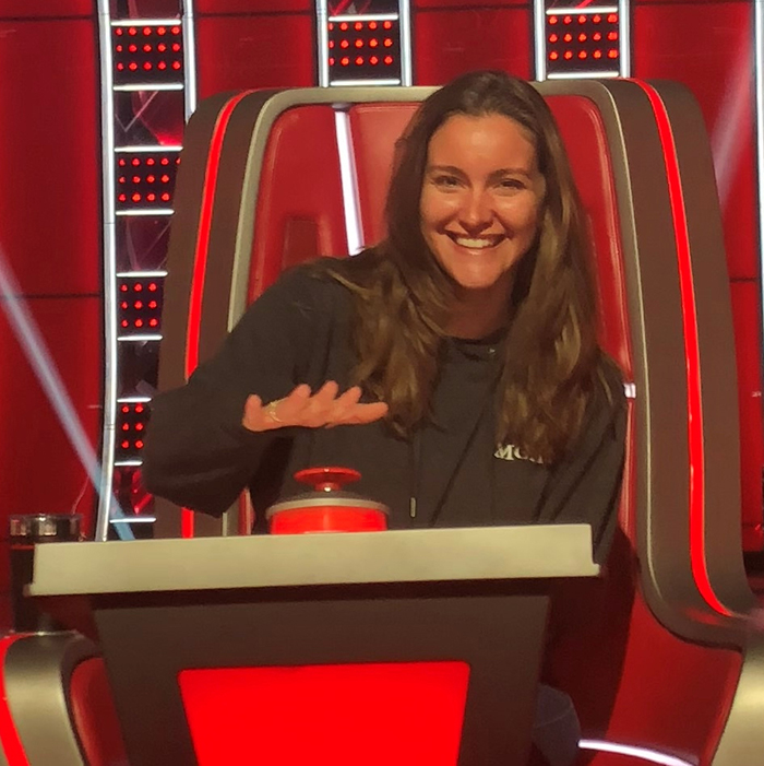 shawna benfield site in a red chair on the set of the Voice, holding her hand over the buzzer