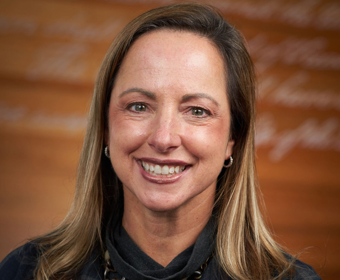 Professor Beth Kubala, a white woman with shoulder-length brown hair, wearing a black and gray plaid jacket over a black blouse with a gold necklace and gold earrings, smiles in front of an orange background.