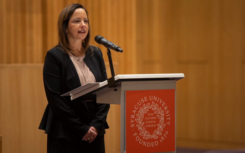 A woman standing at a podium with the Syracuse University logo, delivering a speech in a formal setting. She is wearing a professional outfit with a blazer and light-colored top, speaking into a microphone with wood-paneled walls in the background.