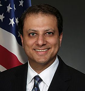 Preet Bharara smiling at camera in a black suit and white button down shirt in front of an american flag