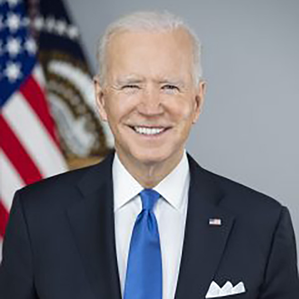 president biden smiling at the camera in a black suit, blue tie, and in front of an american flag