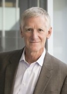 Professor William Banks, a white man white short white hair, wearing a brown suit jacket over a white collared shirt, smiles.