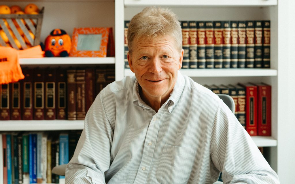 George Boehme looks up from his laptop and smiles. Behind him is a bookshelf.