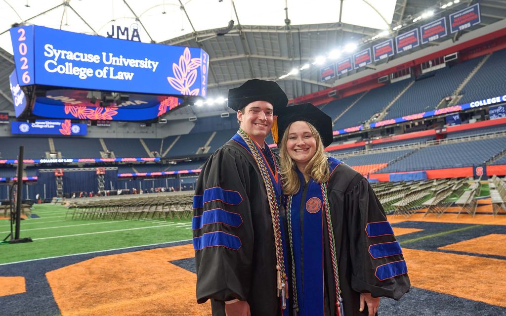 Two graduating students stand on the field in the Syracuse Dome, with a large screen in the background.