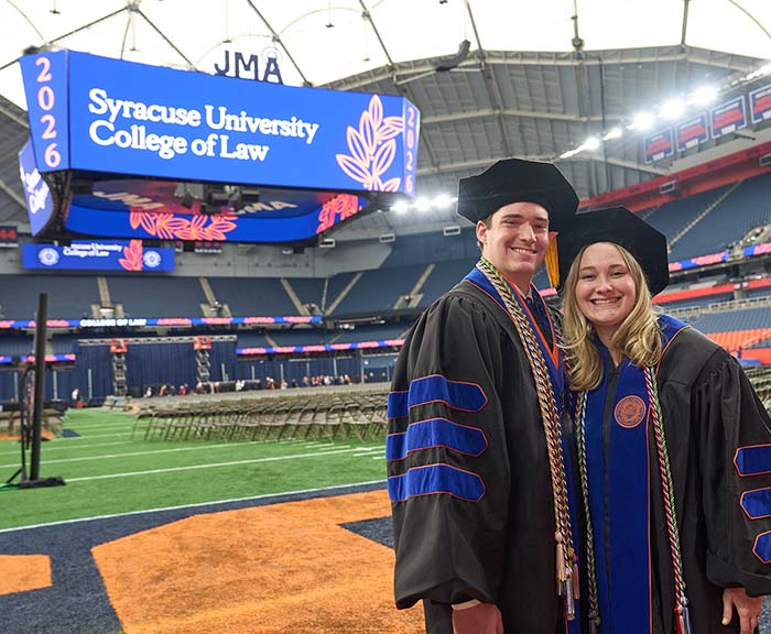 Two graduating students stand on the field in the Syracuse Dome, with a large screen in the background.