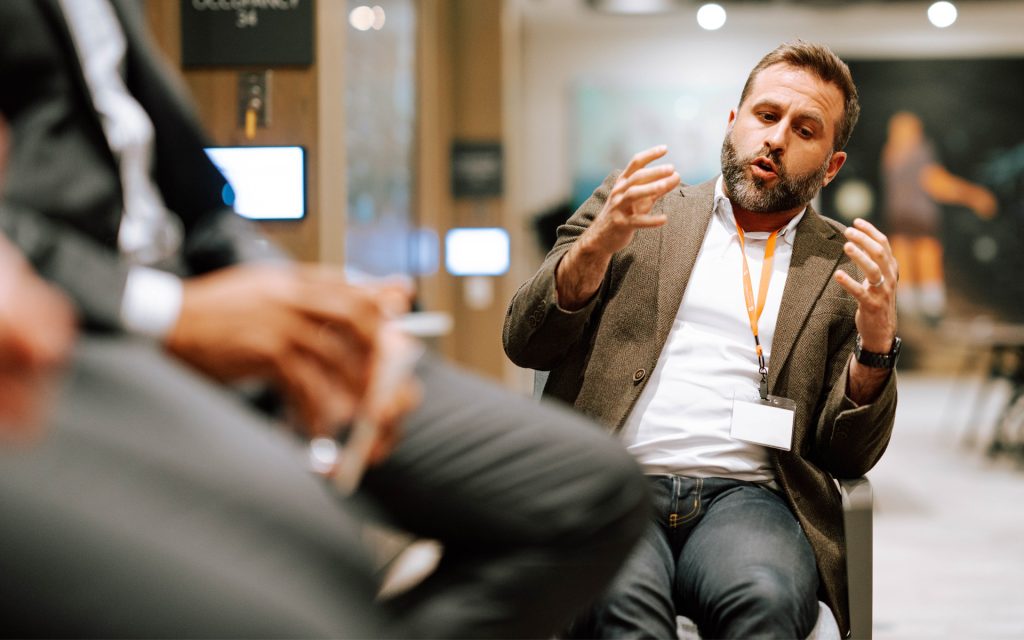 A man wearing a brown blazer and white shirt speaks animatedly, gesturing with his hands while sitting in a professional setting. The background shows a blurred office environment with other seated individuals.