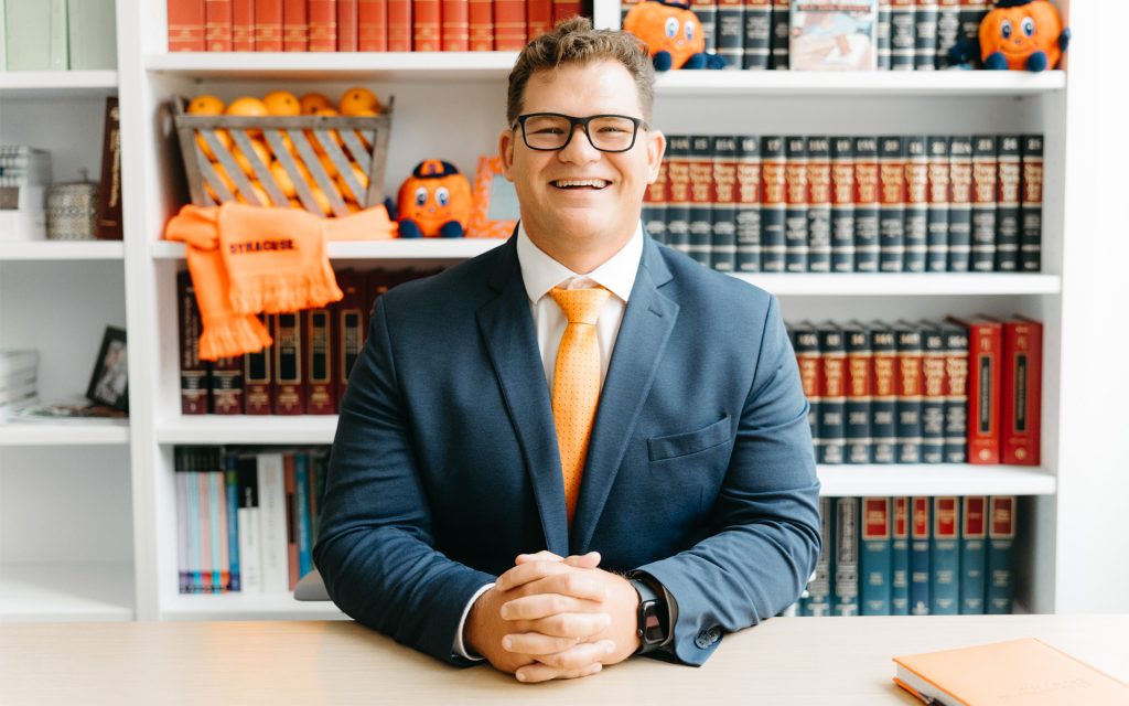 A professionally dressed individual wearing a blue suit, white shirt, and orange tie sits at a desk smiling confidently. Behind them, shelves are filled with law books, Syracuse University-themed items, and a basket of oranges.