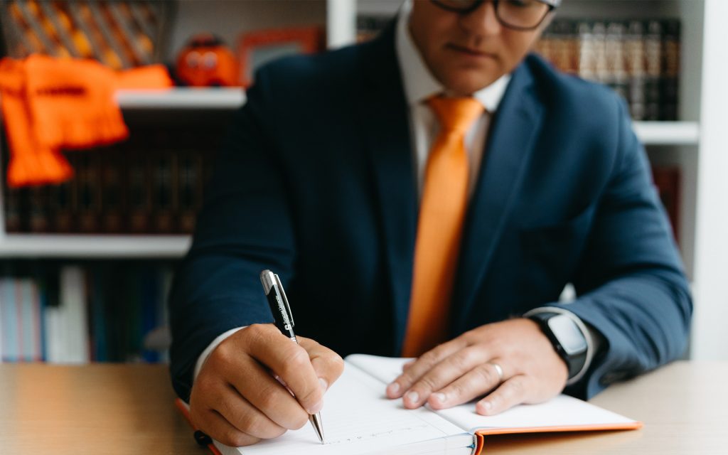 A close-up of the same individual writing in an open notebook on a desk. They wear a blue suit and orange tie, with Syracuse-themed decor and law books visible in the background.