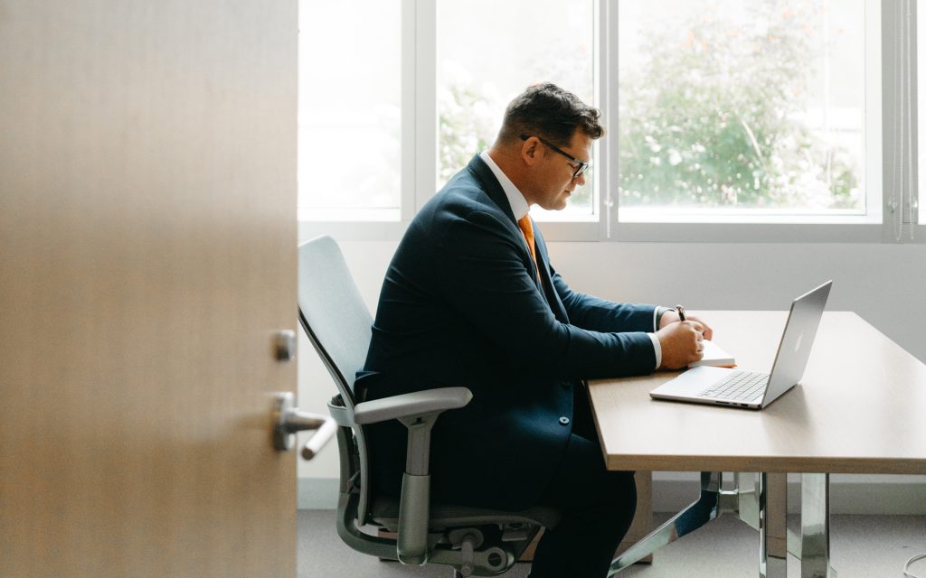 he individual sits at a desk in a well-lit office, focused on writing in a notebook. A laptop is also on the desk, and a window with greenery outside is visible in the background.