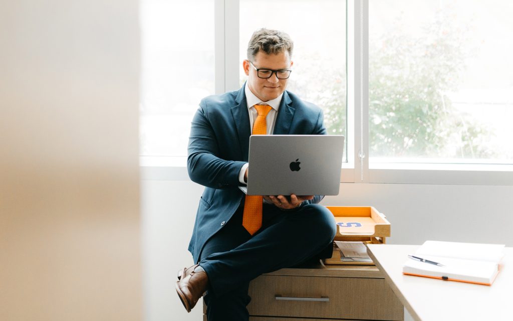 The individual, still in professional attire, sits on the edge of a desk holding a laptop, looking at the screen. The office is bright, with natural light streaming through the windows, and the desk contains Syracuse-themed items and office supplies.