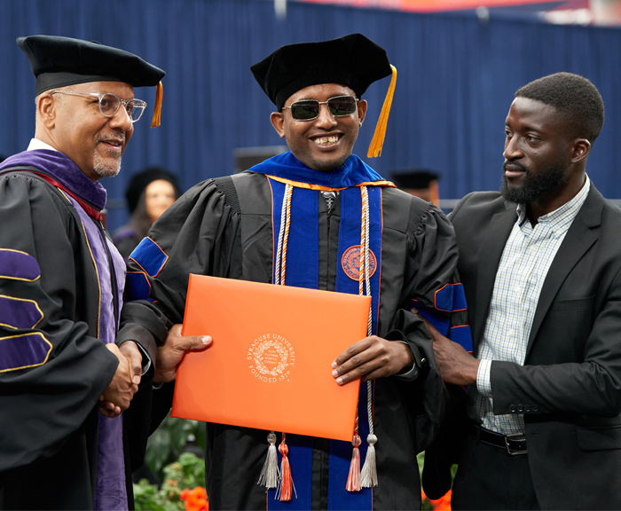 A blind student wearing dark glasses accepts his diploma from the dean of the law school and smiles at the camera.