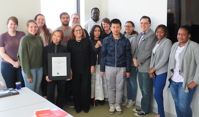 Image Description: Professor Kanter, a white woman with brown should-length hair and glasses wearing black dress pants, a black dress jacket with buttons down the center and pearls stands in the corner of a room with white walls, surrounded by 14 students dressed in casual attire. To Professor Kanter’s immediate right, student Renci Xie a short woman wearing a black zipped coat and black pants, holds the Zero Project Award, a white certificate in a black frame.