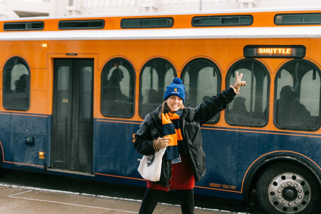student holding up two fingers in front of shuttle, wearing a syracuse hat and scarf