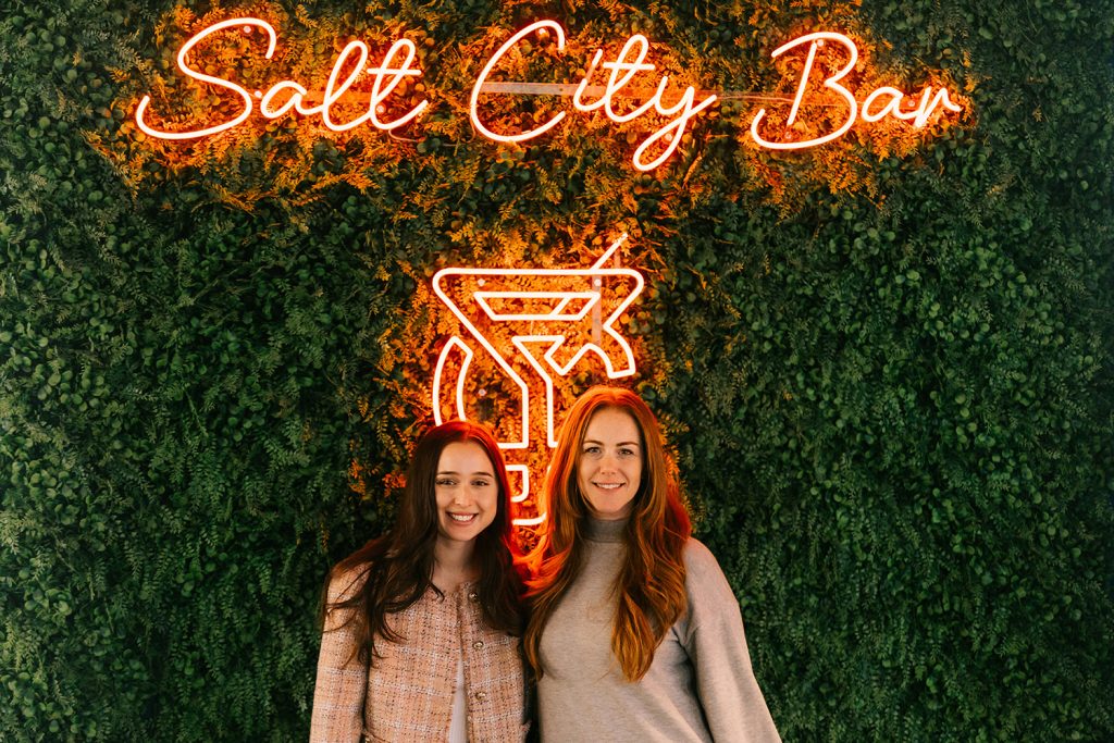 two students standing in front of a salt city bar lit up sign and greenery in the background