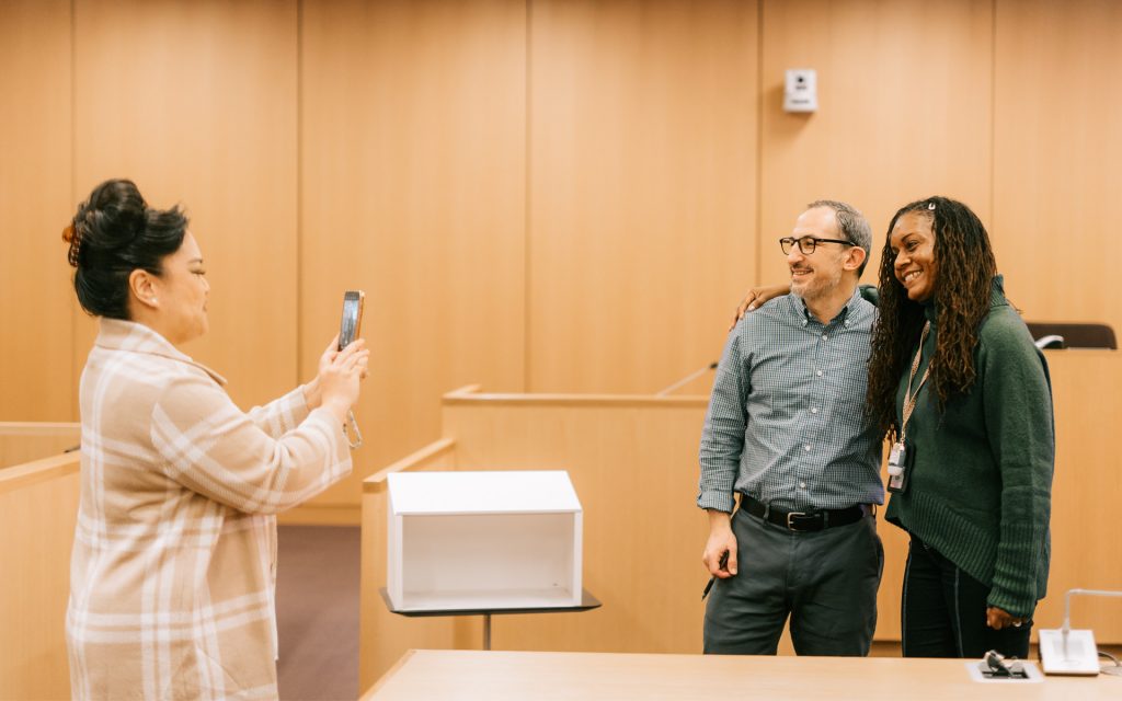 person wearing a plaid coat holds up a phone to take a photo of two individuals posing together in a courtroom setting. The individuals are smiling, with one having their arm around the other. The background shows wooden panels and a podium.