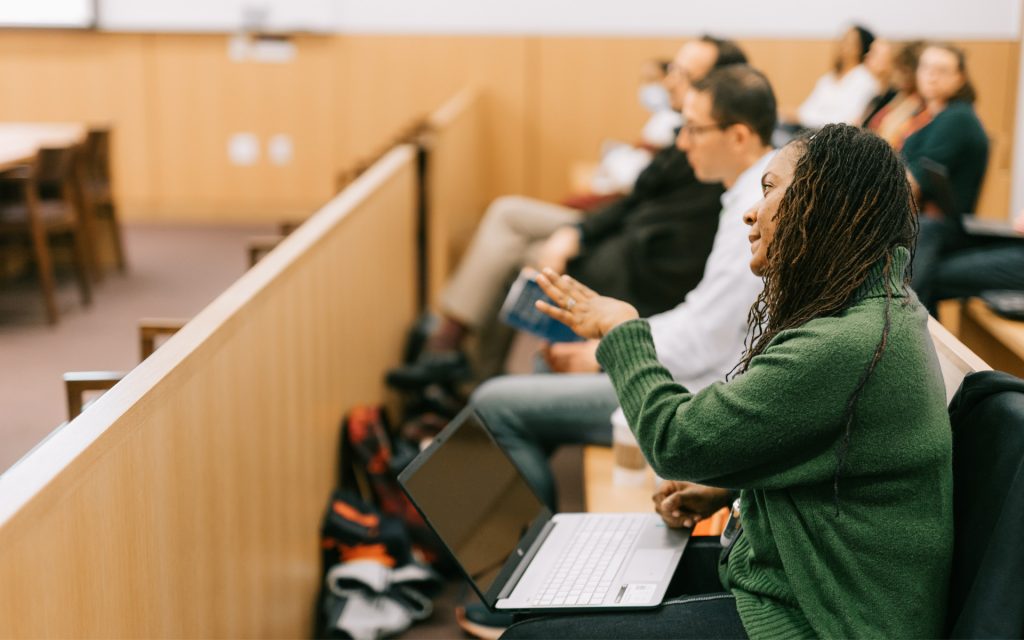 A woman in a green sweater raises her hand to make a comment or ask a question while seated in a lecture hall. She has a laptop open on her lap, and other attendees are seated nearby, listening attentively.