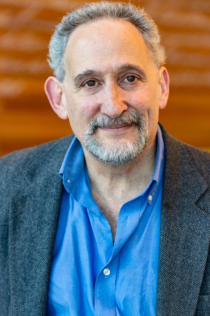 Professor David Driesen, a white man with short gray hair and a gray goatee, wearing a gray suit jacket over a blue collared shirt smiles in front of an orange background.