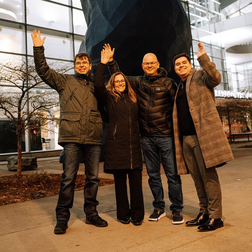 four students smile at the camera and hold their arms up in front of the Denver blue bear statue