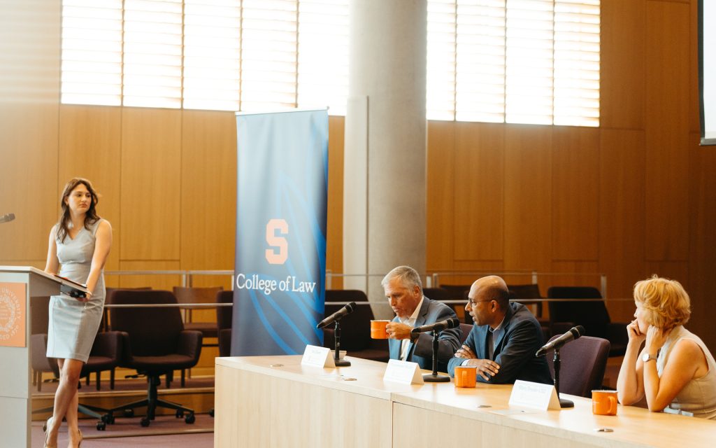 A woman presenting at a podium in a lecture hall with "College of Law" banners. Three panelists are seated at a table with microphones and orange mugs, listening to the presentation.