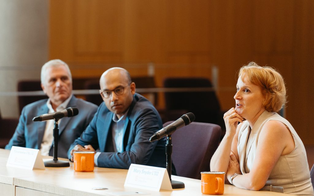 A close-up of a panel discussion in a lecture hall. A woman speaks into a microphone, with two male panelists listening attentively, seated at a table with microphones and orange mugs.