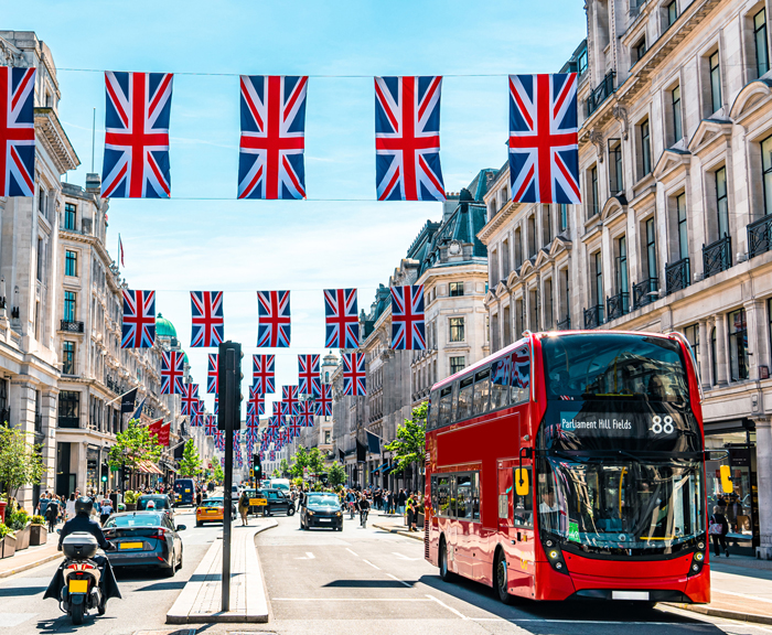 A red double decker bus on a street with British flags hung between the buildings