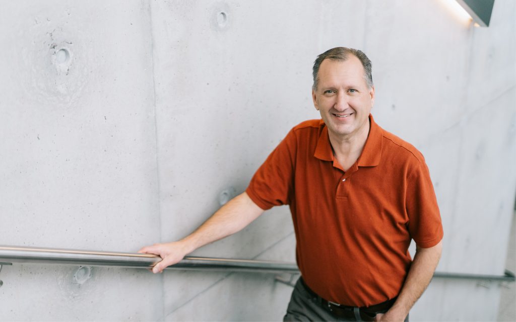 John Dougall stands on the stairs in Dineen Hall and smiles at the camera.