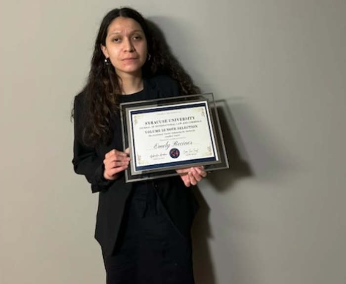 Emely Recinos standing in front of blank wall holding a Syracuse University certificate in a frame.