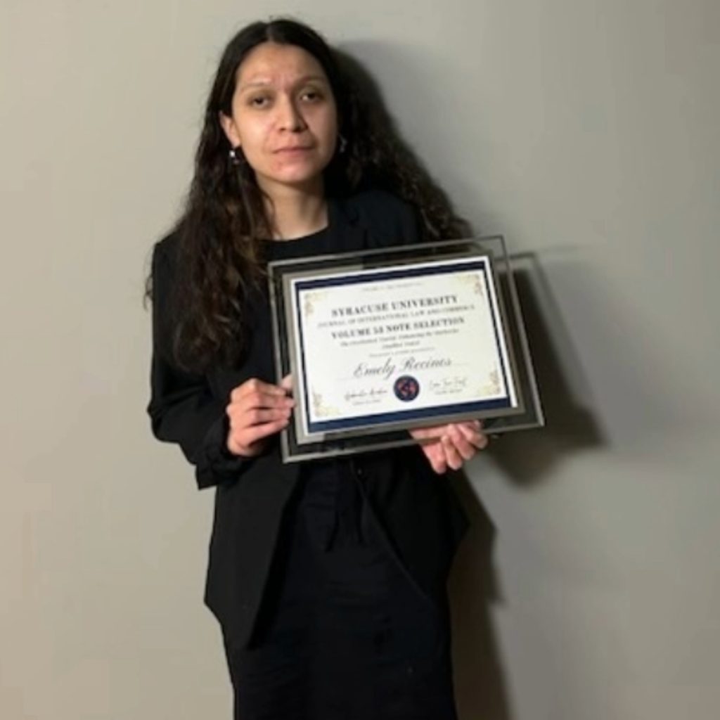 Emely Recinos standing in front of blank wall holding a Syracuse University Certificate in a frame. 