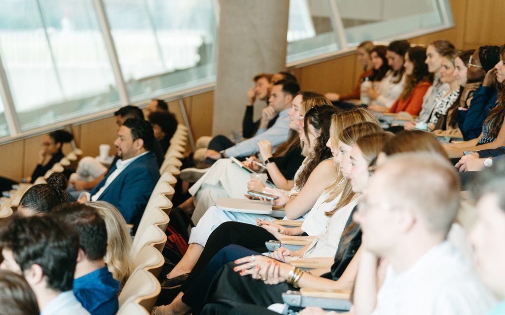 Audience members seated in rows in a brightly lit lecture hall, engaged and taking notes during a presentation.